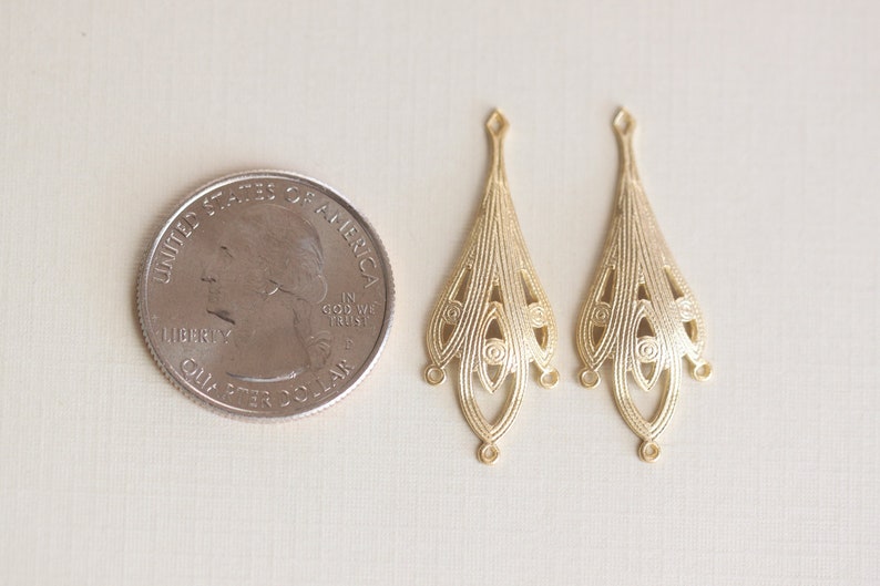May include: Two gold-toned metal earring charms with a delicate, intricate design. The charms are shaped like teardrops with a scalloped edge and a series of concentric circles. The charms are about the size of a US quarter.