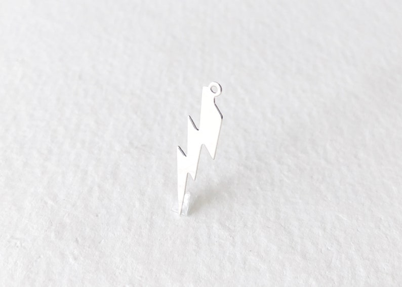 May include: A silver-colored lightning bolt charm with a small loop at the top for attaching to jewelry. The charm has a jagged zig-zag design and is set against a white background.
