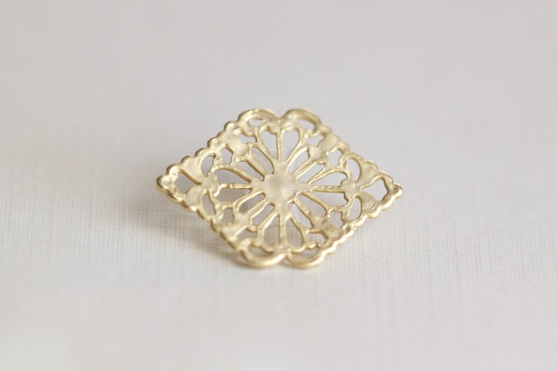 May include: A gold-colored metal decorative piece with a square shape and intricate cut-out design. The design features a floral pattern with a central circle.