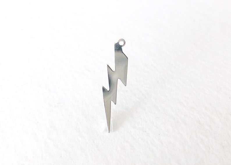 May include: A silver-colored lightning bolt pendant with a small loop at the top for hanging. The pendant has a jagged, zig-zag design, and is displayed against a white background. This jewelry item is suitable for necklaces or earrings.