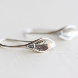 May include: A pair of silver-colored earrings. Each earring features a teardrop-shaped design with a curved hook. The earrings have a smooth, polished finish, reflecting light. The earrings are likely made of sterling silver or a similar metal.