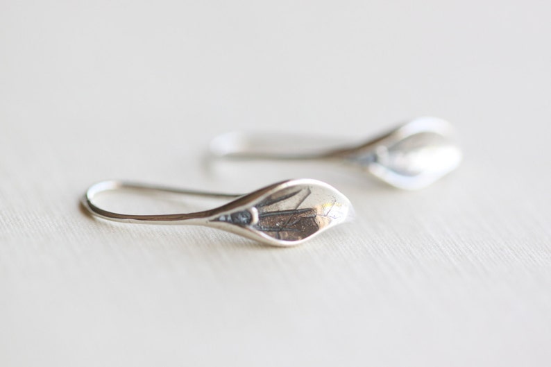 May include: A pair of silver-colored earrings. Each earring features a teardrop-shaped design with a curved hook. The earrings have a smooth, polished finish, reflecting light. The earrings are likely made of sterling silver or a similar metal.