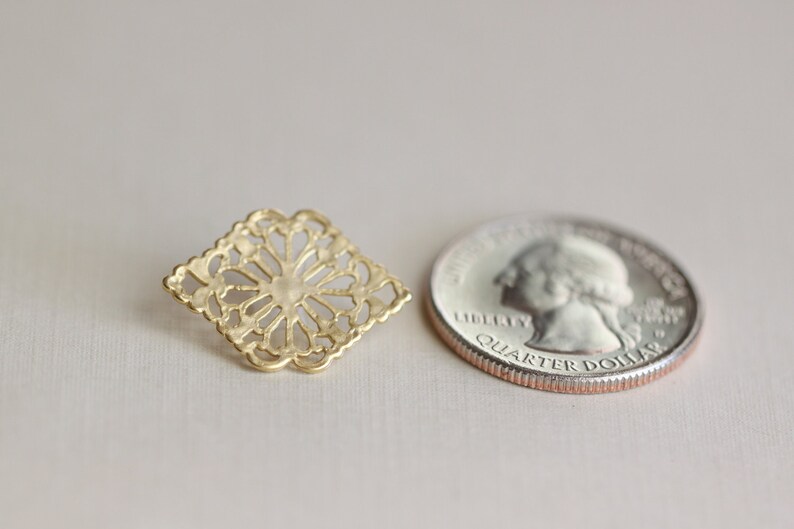 May include: A gold-colored metal charm with a square, intricate, cutout design. The charm is about the size of a US quarter.