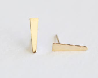 May include: A pair of gold-colored stud earrings. Each earring has a simple, geometric design, with a flat, elongated triangle shape. The earrings are displayed on a white background.