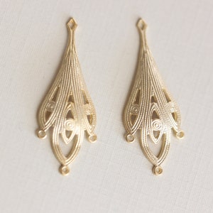 May include: Two gold-toned metal earring charms with a delicate, intricate design. Each charm features a teardrop shape with multiple layers of openwork.