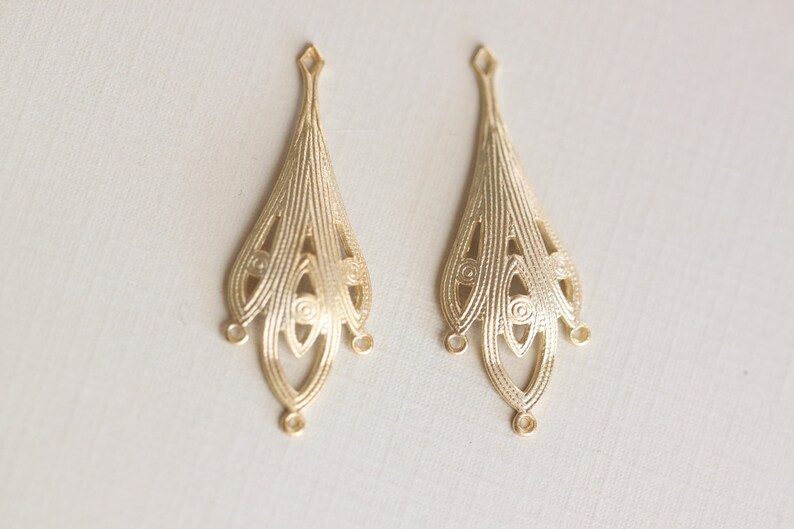 May include: Two gold-toned metal earring charms with a delicate, intricate design. Each charm features a teardrop shape with multiple layers of openwork.