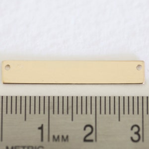 May include: A gold-colored metal bar with two holes, measuring approximately 3 cm in length. The bar is shown next to a ruler for scale.