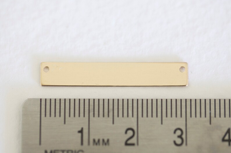 May include: A gold-colored metal bar with two holes, measuring approximately 3 cm in length. The bar is shown next to a ruler for scale.