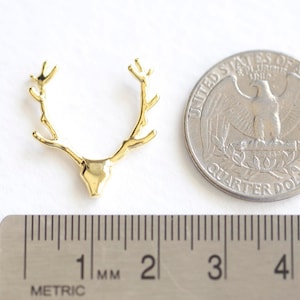 May include: A gold-colored deer antler pendant, next to a US quarter coin and a metric ruler. The pendant is approximately 1 inch wide. The quarter coin shows an eagle. The ruler shows measurements in millimeters.