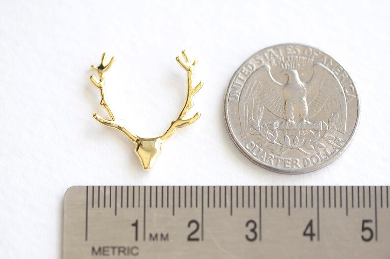 May include: A gold-colored deer antler pendant, next to a US quarter coin and a metric ruler. The pendant is approximately 1 inch wide. The quarter coin shows an eagle. The ruler shows measurements in millimeters.