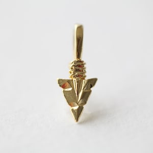 May include: A gold-colored arrowhead pendant with a detailed, faceted design. The pendant has a loop at the top for attaching to a chain or cord.