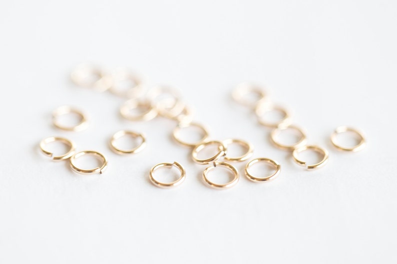 May include: A group of small, gold jump rings, scattered on a white surface. These rings are used for jewelry making and crafting.