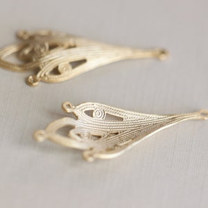 May include: Two gold-colored metal filigree embellishments with intricate designs. The embellishments are shaped like teardrops with a pointed end and a rounded top.