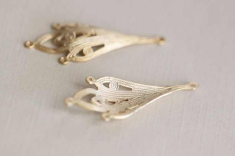May include: Two gold-colored metal filigree embellishments with intricate designs. The embellishments are shaped like teardrops with a pointed end and a rounded top.