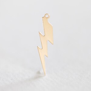 May include: A gold-colored lightning bolt charm with a small loop at the top for hanging. The charm is a simple, stylized design, perfect for jewelry making or other craft projects. The charm is approximately 1.5 inches in length.