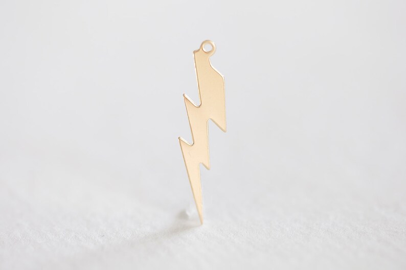 May include: A gold-colored lightning bolt charm with a small loop at the top for hanging. The charm is a simple, stylized design, perfect for jewelry making or other craft projects. The charm is approximately 1.5 inches in length.