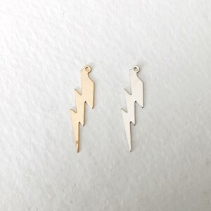 May include: Two metal lightning bolt charms, one gold-colored and one silver-colored, are displayed against a white background. Each charm has a small loop at the top for attaching to a necklace or bracelet. The lightning bolt design is a popular symbol.