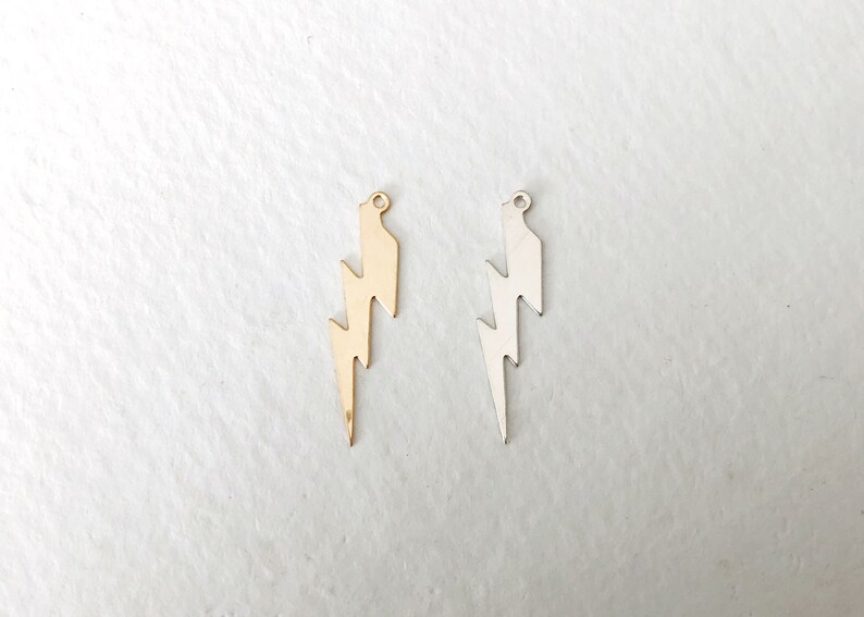 May include: Two metal lightning bolt charms, one gold-colored and one silver-colored, are displayed against a white background. Each charm has a small loop at the top for attaching to a necklace or bracelet. The lightning bolt design is a popular symbol.