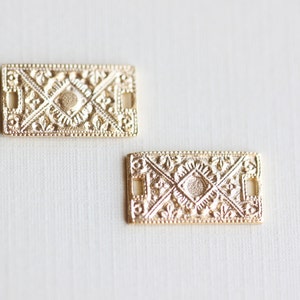 Puede incluir: Dos adornos rectangulares de metal dorado con intrincados diseños florales y geométricos. Los adornos tienen dos pequeños agujeros a cada lado.