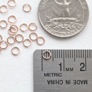 May include: A close-up of a pile of 20 small, rose gold jump rings, measuring 1 mm in diameter, next to a US quarter and a ruler.  The jump rings are used for jewelry making.