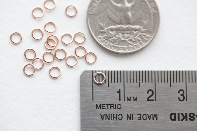May include: A close-up of a pile of 20 small, rose gold jump rings, measuring 1 mm in diameter, next to a US quarter and a ruler.  The jump rings are used for jewelry making.