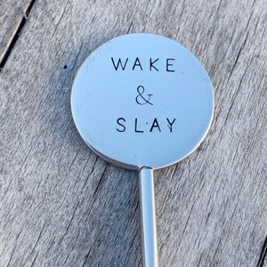 May include: A silver metal stirring spoon with a round, flat end that says "WAKE & SLAY" in black lettering.