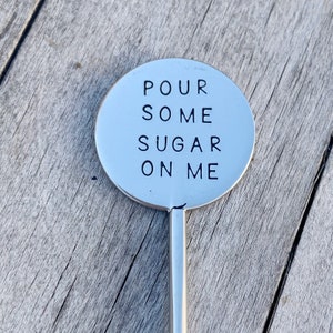 May include: A silver-colored, round cocktail stirrer with the words "POUR SOME SUGAR ON ME" in black, centered on the round top. The stirrer has a long, thin handle and is set against a weathered wooden surface.