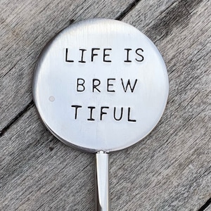 May include: A silver-colored, round bottle stopper with the words "LIFE IS BREWTIFUL" stamped in black, centered on the disc. The stopper has a long, thin handle. The background is a wooden surface.