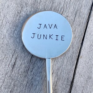 May include: A silver stirring spoon with a round, flat end that says "JAVA JUNKIE" in black lettering.