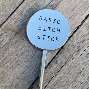 May include: A silver metal stirring stick with a round, flat top that says "BASIC BITCH STICK" in black lettering.