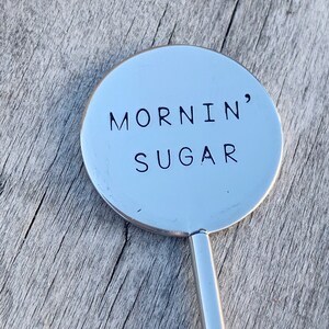 Puede incluir: Una cuchara de revolver de plata con una parte superior redonda y plana que dice "MORNIN' SUGAR" en letras negras.