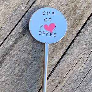 May include: A silver stirring stick with a round, silver disc at the top. The disc has the text "CUP OF OFFEE" with a pink heart replacing the "O" in "OFFEE".