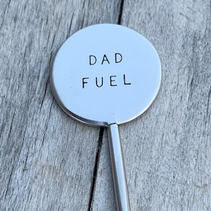 May include: A silver metal stirring spoon with a round, flat end that says "DAD FUEL" in black lettering.