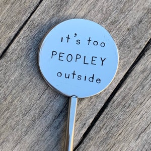 May include: A silver metal cocktail stirrer with a circular top. The top has the stamped text "it's too PEOPLEY outside" in a simple, sans-serif font. The stirrer is set against a weathered wooden surface.