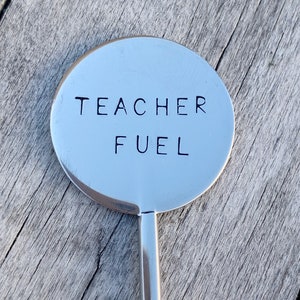 May include: A silver metal stirring spoon with a round handle that says "TEACHER FUEL" in black lettering.