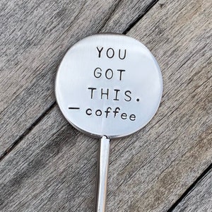 May include: A silver-colored coffee scoop with a circular head. The head of the scoop has the words "YOU GOT THIS. - coffee" stamped on it in a bold, sans-serif font. The scoop is set against a wooden background.
