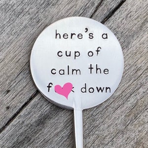 May include: A silver metal stirring spoon with a round handle that says "Here's a cup of calm the f**k down" with a pink heart replacing the "k".