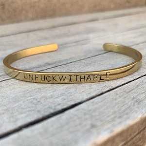 May include: Gold-toned metal bracelet with the words "Unfuckwithable" engraved on the band.