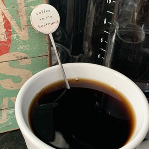 May include: A silver coffee stirrer with a round charm that says "coffee is my boyfriend". The stirrer is in a white mug with black coffee.