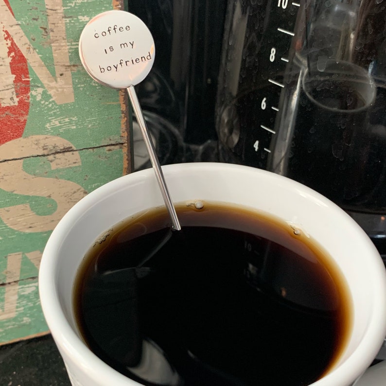 May include: A silver coffee stirrer with a round charm that says "coffee is my boyfriend". The stirrer is in a white mug with black coffee.