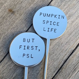 May include: Two silver metal signs with black text. The top sign reads "Pumpkin Spice Life". The bottom sign reads "But First, PSL".