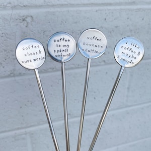 May include: Four silver metal stirring sticks with round metal tops. Each top has a different saying about coffee. The sayings are: "coffee chaos & cuss words", "coffee is my spirit animal", "coffee because adulting", and "i like coffee & maybe 3 people".