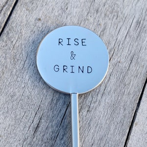 May include: A silver metal sign with the words "RISE & GRIND" engraved on it. The sign is attached to a silver metal stick.