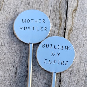 May include: Two silver metal stirring sticks with round ends. The top of each stick has a phrase engraved on it. The first stick says "MOTHER HUSTLER". The second stick says "BUILDING MY EMPIRE".