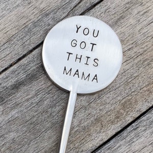 May include: A silver metal plant marker with the words "YOU GOT THIS MAMA" stamped on a circular head. The marker has a long, thin stem and is set against a weathered wooden surface.