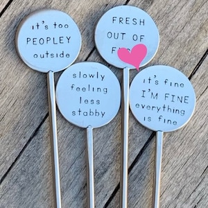 May include: Four silver plant markers with humorous phrases. The markers have round, light blue faces with black text. Phrases include "it's too PEOPLEY outside," "FRESH OUT OF F," "slowly feeling less stabby," and "it's fine I'M FINE everything is fine."