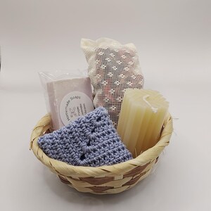 May include: A small woven basket containing a bar of soap in clear plastic packaging with a label reading Homemade Soaps Purple Lavender Glycerin, a light blue crocheted washcloth, a white mesh bag, and a ribbed off-white pillar candle.