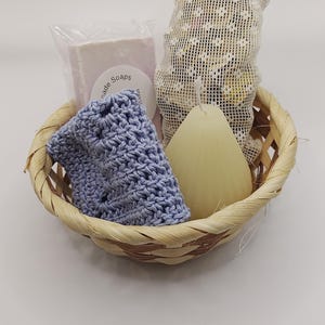 May include: A woven wicker basket containing a bar of soap in clear plastic packaging with a label reading Handmade Soaps, a light blue crocheted washcloth, a cream-colored teardrop-shaped candle, and a white mesh bag filled with small items.