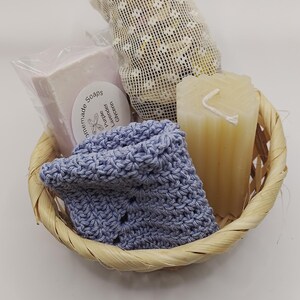 May include: A light-colored woven basket containing a bar of purple lavender glycerin soap in clear plastic wrap, a beige pillar candle, a small white mesh drawstring bag, and a folded periwinkle blue crocheted square cloth item.