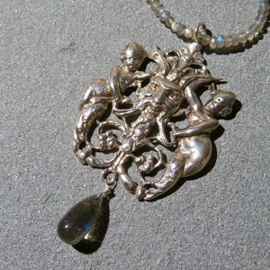 May include: A silver pendant necklace featuring an intricate design of a face surrounded by two figures. The pendant is attached to a silver chain with a teardrop-shaped gemstone.